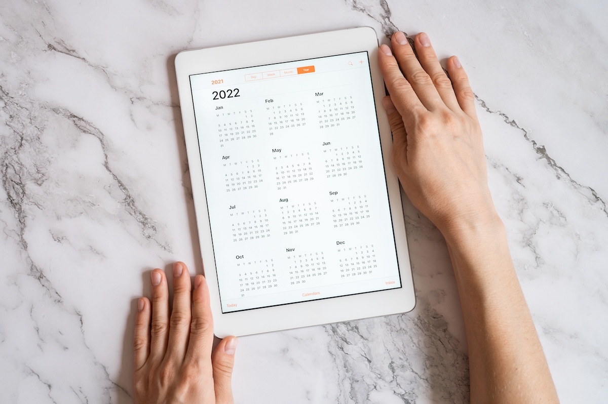 Tablet computer with an open app of calendar for 2022 year in a womans hands on a gray marble background. concept business or to do list goals with technology using. top view, flat lay